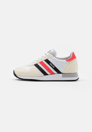 Biały sneaker z czarnymi i czerwonymi paskami, beżowymi zamszowymi akcentami, czerwoną zakładką na pięcie i napisem "USA 84" z boku, na białym tle.