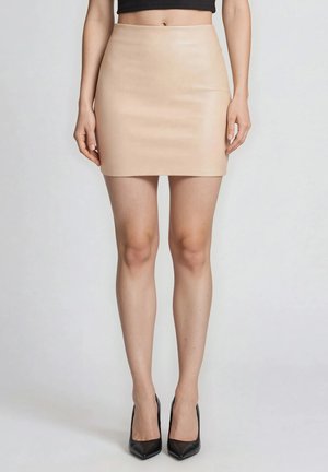 Nainen, jolla on beige minihame, musta crop top ja mustat terävät korkokengät, seisoo yksiväristä vaaleaa taustaa vasten.