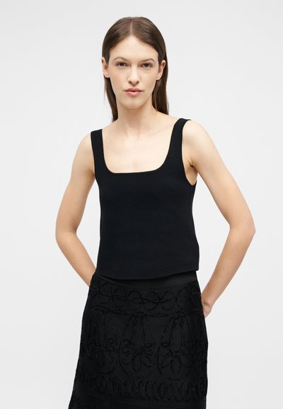 Femme aux cheveux bruns lisses portant un haut sans manches noir à encolure carrée et une jupe noire avec un motif floral brodé, mains derrière le dos.