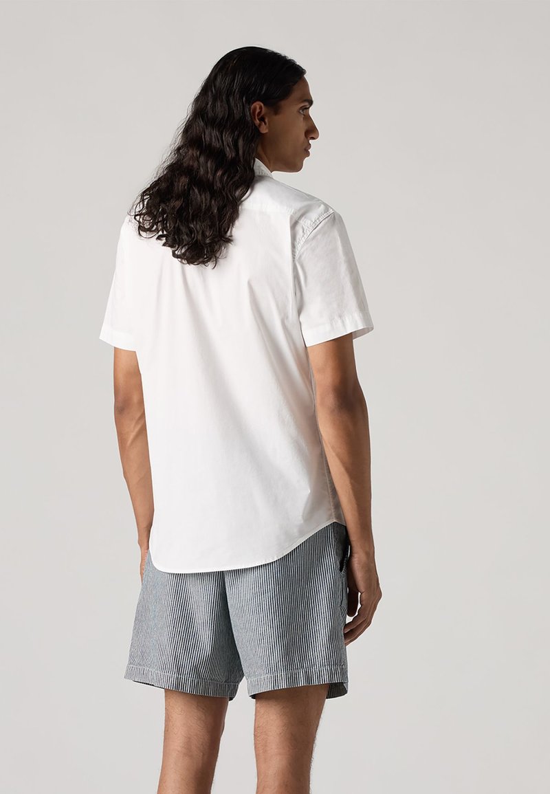 Chemise blanche à manches courtes en tissu coton, coupe droite, détail de l'empiècement au dos, assortie à un short gris rayé avec poche latérale visible.