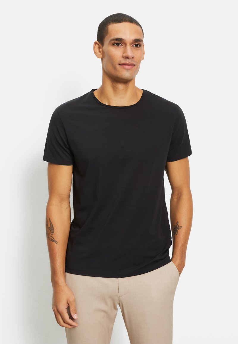T-shirt nera a maniche corte con scollatura rotonda, realizzata in tessuto di cotone liscio, indossata da una persona con pantaloni beige.