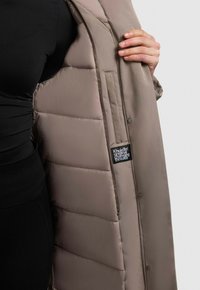 Svetložltý pufový kabát s prešívaným vzorom, s bočnou kapsou a štítkom s nápisom "Khujo Original Changing Threads."