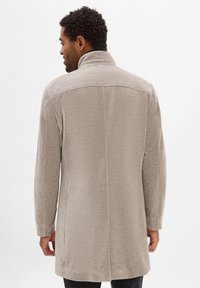 Mężczyzna stojący tyłem, ubrany w jasnobeżowy płaszcz do kolan z wysokim kołnierzem i długimi rękawami, na jednolitym białym tle.