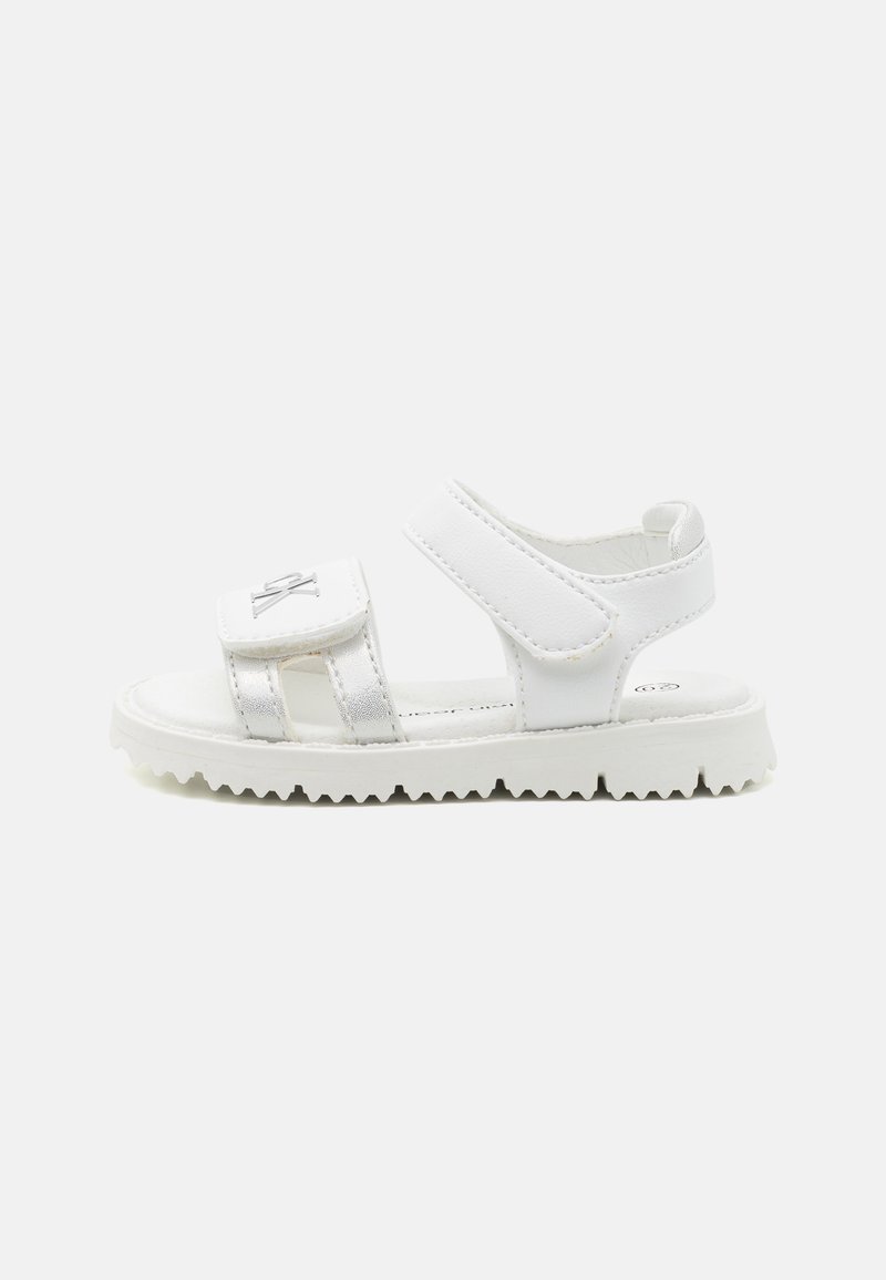 White sandals featuring a velcro strap and a silver accent. Designed with a textured sole and a flat profile for comfort.