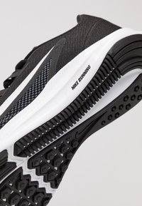 Chaussure de course noire en synthétique avec un dessus en maille texturée, semelle intermédiaire blanche contrastante et semelle en caoutchouc présentant des motifs hexagonaux.