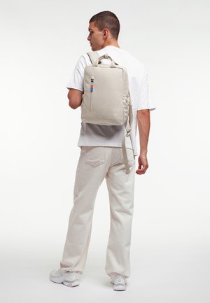 Joven con camiseta blanca, pantalones beige claro y zapatillas blancas, lleva una mochila rectangular beige claro colgada de un hombro.