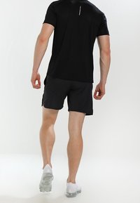 Czarna sportowa koszulka z krótkim rękawem z odblaskowym szwem z tyłu, w zestawie z ciemnymi szortami i białymi trampkami z przezroczystą podeszwą.