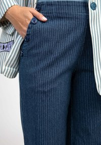 Pantalon bleu marine à fines rayures en denim, avec poches latérales et boutons bleus le long de la ceinture. Associé à une chemise rayée.