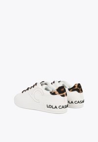 Bijele tenisice s leopard uzorkom na peti, kontrastnim vezicama i oznakom 'LOLA CASADA' na stražnjem dijelu. Glatki materijal, zaobljen oblik.