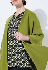 Zelený pletený cardigan přes námořnický top s vzorem, který obsahuje bílé a zelené vlnité čáry. Doplňky zahrnují zlaté kroužkové náušnice.