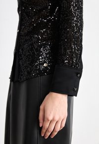 Zwarte paillettenjas met een strak ontwerp, voorzien van een geruite manchet en een gladde textuur. Accenten zijn glimmende knopen tegen een matte stof.