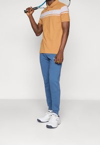 Pomarańczowa koszulka polo w białe paski, niebieskie dresy i białe buty sportowe. Zaprojektowana do sportu, z kołnierzykiem i detalem logo.