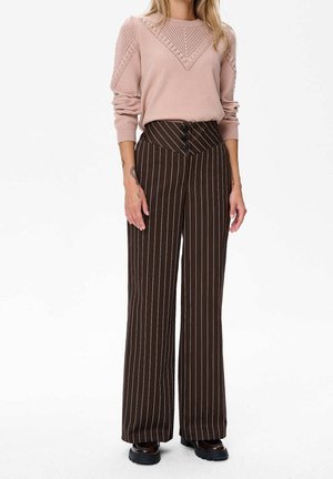 Frau trägt hellrosa langärmeligen Strickpullover und hoch taillierte braune Nadelstreifenweite Hose mit schwarzen Schuhen, stehend auf weißem Hintergrund.