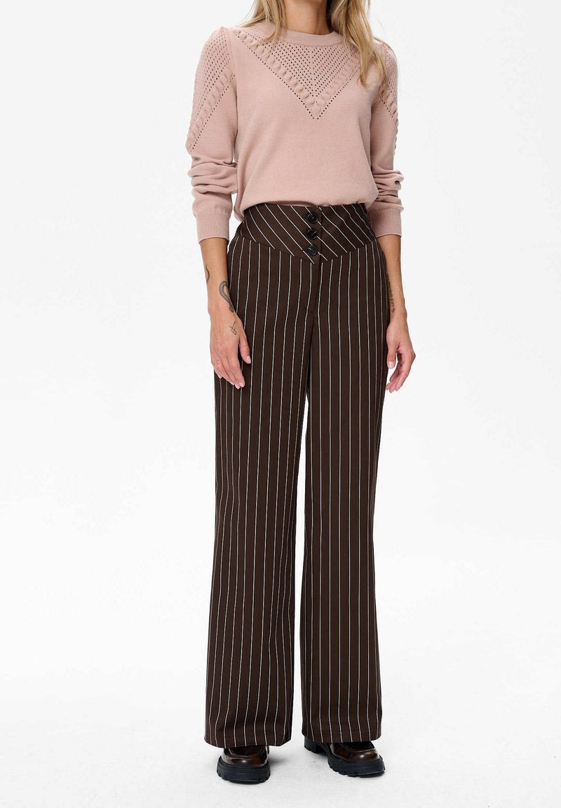 Femme portant un pull en maille rose clair à manches longues et un pantalon marron à rayures larges taille haute avec des chaussures noires, debout sur fond blanc.