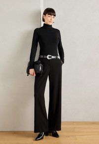 Top negro de cuello alto combinado con pantalones anchos negros, acentuado por un cinturón plateado con tachuelas y un bolso negro pequeño. Zapatos de tacón negros.