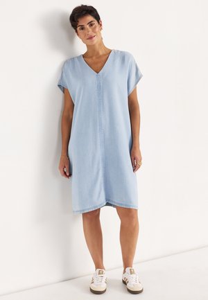 Frau mit kurzen dunklen Haaren, die ein hellblaues, knielanges Kleid mit V-Ausschnitt und weiße Turnschuhe trägt, steht vor einer weißen Wand.