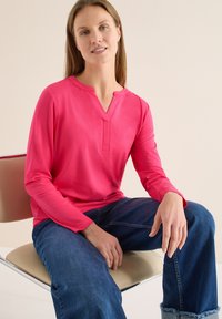 Haut rose à manches longues avec encolure en V et détails de boutons, associé à un jean bleu. Tissu doux, coupe décontractée, design minimaliste.