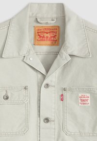 Camisa de mezclilla gris claro con un cuello clásico, dos bolsillos frontales, botones a presión plateados y una etiqueta roja de Levi's. La etiqueta dice "LEVI STRAUSS & CO."
