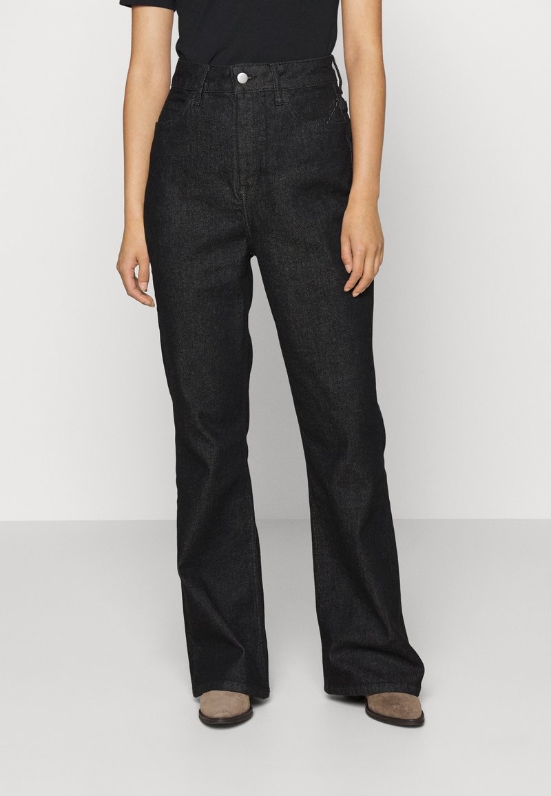 Personne portant un jean denim noir taille haute et des chaussures beige, debout devant un fond clair uni.