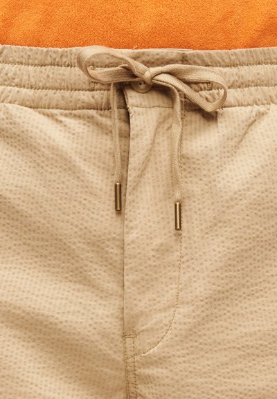 Μπεζ σορτς με υφή, με κορδόνι στη μέση που δένει και κλείσιμο με κουμπί, πάνω σε πορτοκαλί υφασμάτινο φόντο.
