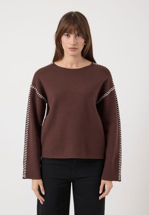 Frau in einem braunen Langarmpullover mit weißen gestickten Details an den Ärmeln und Schultern, kombiniert mit schwarzen Hosen, die nach vorne schaut.