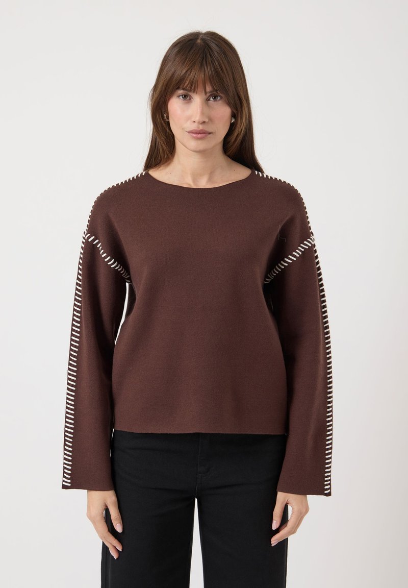 Femme portant un pull long à manches longues marron avec des détails en fil blanc le long des manches et des épaules, associé à un pantalon noir, regardant vers l'avant.