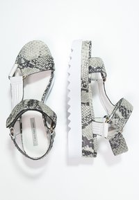 Dámské platformové sandály s bílými tlustými zigzagovými podešvemi a šedými hadími popruhy, kterými se dají nastavit bílé kotníkové popruhy.