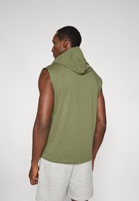 Φουτερ με κουκούλα χωρίς μανίκια σε ελιά, με μεγάλη κουκούλα, κατασκευασμένο από απαλό ύφασμα. Φορεμένο με ανοιχτό γκρι σορτς, δείχνοντας ένα casual αθλητικό στυλ.