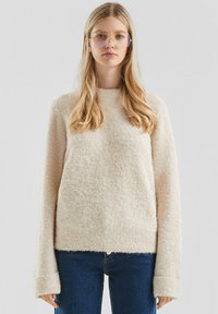 Młoda kobieta z długimi blond włosami nosi oversize'owe okulary, beżowy sweter o teksturze i niebieskie dżinsy na jednolitym tle.