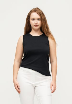 Vrouw met lang rood haar, gekleed in een zwart mouwloos shirt en witte broek, staand tegen een effen lichte achtergrond.