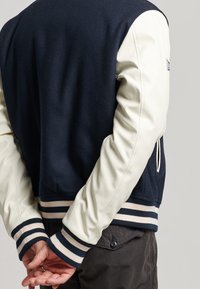 Marinblått ullmix-jacka med krämfärgade läderslevar, randiga ribbmuddar och elastiskt midjeband. Detaljerad sömnad synlig vid sömmarna.