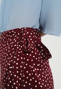 Burgundská sukně s puntíky a uvázanou mašlí v pase, vyrobená z lehkého materiálu. Spárovaná s světle modrým topem s krátkým rukávem.