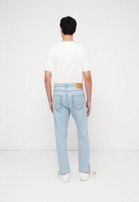 Άνδρας που στέκεται πλάτη, φοράει λευκό μπλουζάκι, ανοιχτόχρωμα μπλε τζιν με πίσω τσέπες και λευκά αθλητικά παπούτσια σε λευκό φόντο.