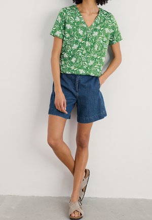 Top corto verde a fiori con maniche corte e scollatura a V, abbinato a pantaloni corti in denim blu. Sandali piatti di colore chiaro completano l'outfit.