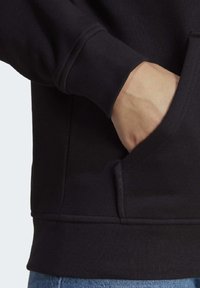 Schwarzer Hoodie mit gerippten Bündchen und einer Vordertasche. Der Stoff scheint weich mit glatter Textur zu sein, teilweise in blaue Jeans gesteckt.
