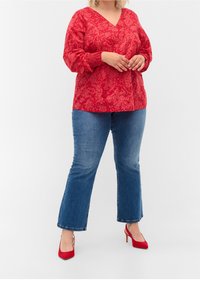 Blusa a V con motivo rosso e maniche a sbuffo lunghe, abbinata a jeans svasati blu e scarpe con tacco rosso. Tessuto testurizzato e design dettagliato.