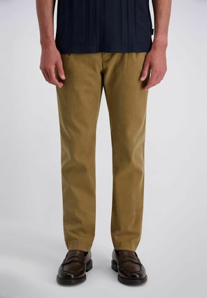 Mann trägt braune, gerade geschnittene Hosen, dunkelbraune Lederloafers und ein dunkles Hemd, sichtbar von der Taille abwärts.