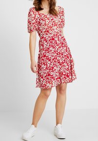 Rotes Blumenkleid mit weißen Blumen, kurzen Puffärmeln, tailliertem Schnitt und knieumspieltem Rock. Kombiniert mit weißen Sneakers.