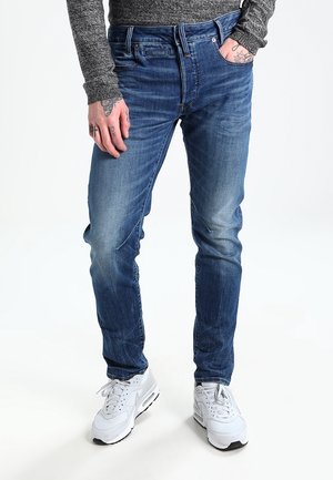 Homme portant un jean bleu, une chemise grise à manches longues et des baskets blanches, avec des tatouages visibles sur les mains, debout avec une main dans la poche.