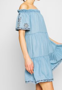 Nainen, joka on pukeutunut vaaleansiniseen olkaimettomaan mekkoon, jossa on brodeeratut lyhyet hihat ja helma, pitää kiinni liehuvan hameen sivusta.