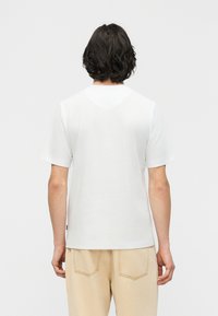 Άνδρας με μεσαίου μήκους σκούρα μαλλιά που φοράει απλό λευκό κοντομάνικο μπλουζάκι και μπεζ παντελόνι, εμφανιζόμενος από πίσω με φόντο λευκό.