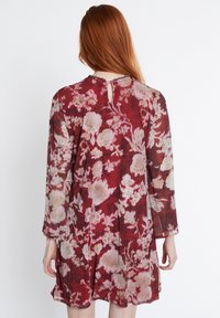 Durchsichtiges Blumenkleid mit rotem Grundton und großen cremefarbenen Blumen, langen Ärmeln und einem Schlüsselloch-Detail am hinteren Hals.