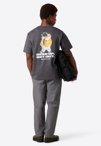 Mand står med ryggen til iført en mørkegrå t-shirt med en tegneserie-bjørn i en orange jakke og teksten "GÅ I GANG. KOM UD." mens han holder en sort taske.