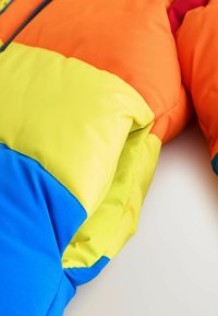 Giacca a vento colorata con sezioni di arancione, giallo e blu. Il materiale è morbido con imbottitura per isolamento e comfort.