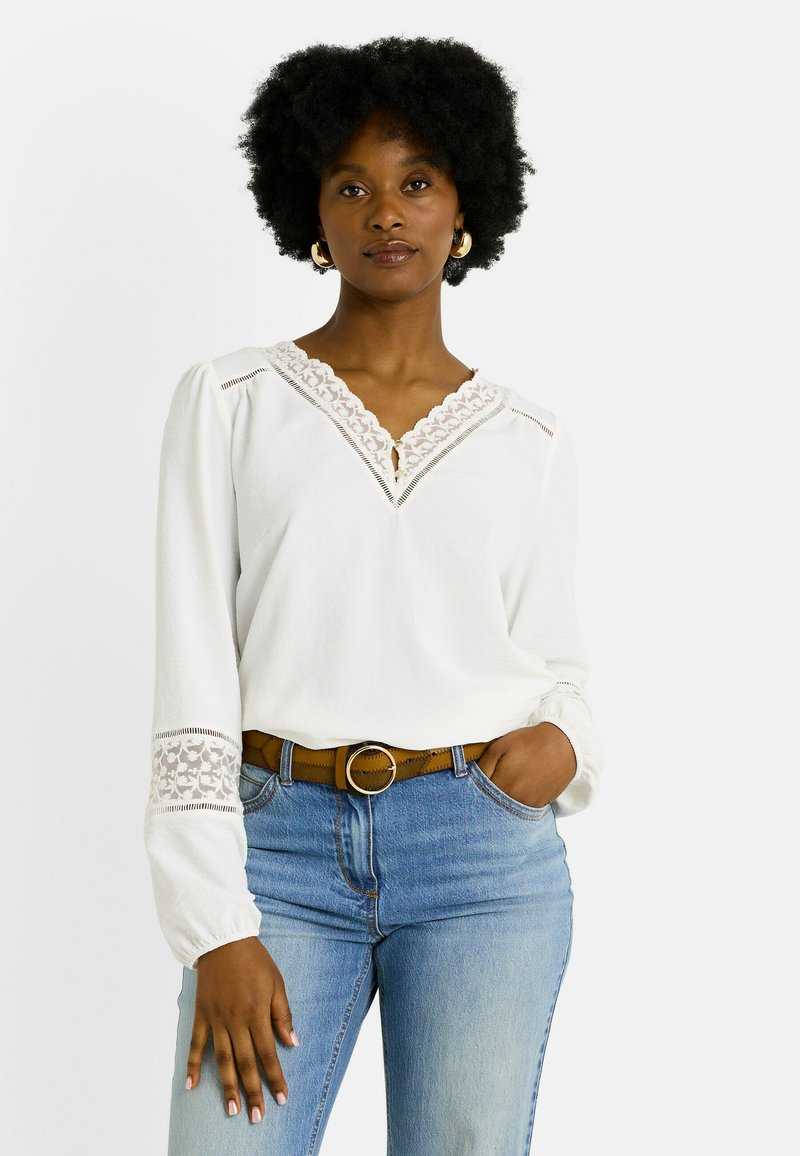 Blusa blanca con encaje en el cuello y las mangas, combinada con jeans de lavado medio y un cinturón marrón con hebilla circular.