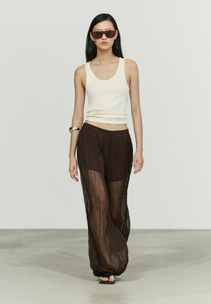 Giovane donna con lunghi capelli neri, che indossa occhiali da sole, un top bianco senza maniche, pantaloni neri larghi e trasparenti, sandali neri e un braccialetto d'argento, cammina avanti.