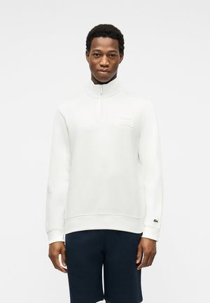 Jeune homme portant un sweat-shirt blanc à fermeture éclair quart et un short foncé, debout devant un fond clair uni, face à l'avant.