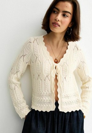 Kobieta ubrana w kremowy, teksturowany sweter z dzianiny z falistymi krawędziami, zawiązany z przodu, w połączeniu z ciemną, wysoką spódnicą.