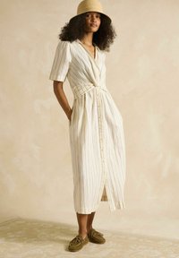 Svītrota linu kleita krēmkrāsā ar bežām līnijām. Pogu aizdare priekšā, jostas saite, īsas piedurknes un kabatas. Valkāta ar olīvzaļiem apaviem.