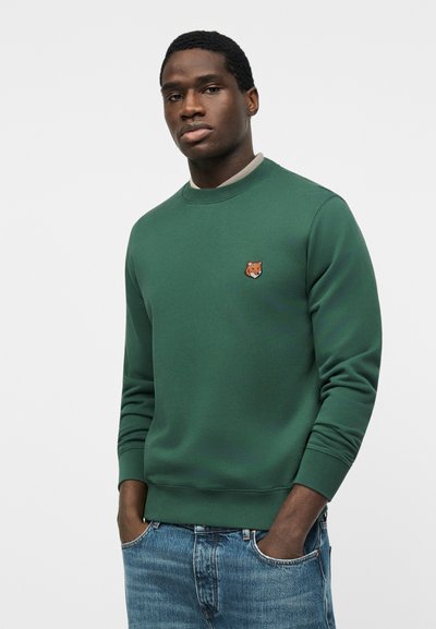 Νεαρός άνδρας που φοράει πράσινο φούτερ με λογότυπο αλεπούς στο στήθος και μπλε τζιν, στέκεται με τα χέρια στις τσέπες μπροστά σε απλό φόντο.